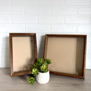 May include: Two wooden picture frames with a brown finish, one rectangular and one square, are displayed. A small white textured pot with green succulents sits in front of the frames. The background is a white brick wall.