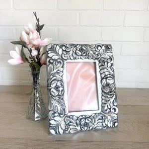 May include: A rectangular picture frame with a metallic silver finish and a raised, ornate floral relief pattern surrounding a central opening. The frame features a smooth inner border and a textured, sculpted outer edge with flower motifs.
