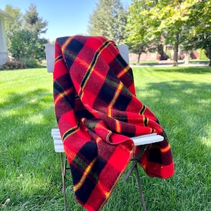 May include: A red and black plaid blanket draped over a white chair, set on a grassy lawn. The blanket features a bold pattern with yellow and green accents. The scene is outdoors on a sunny day.