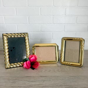 May include: Three gold-toned metal picture frames. Two frames feature a twisted rope-style border, while the center frame has a smooth, beveled edge. A cluster of three vibrant pink artificial flowers with green leaves rests in front of the frames.