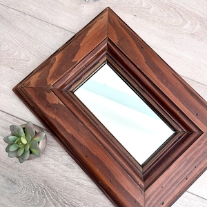 May include: A diamond-shaped mirror with a dark brown wooden frame. The frame has a beveled edge and a natural wood grain pattern. A small, silver-colored pot with a green succulent plant is in the lower left corner.