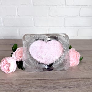 May include: A clear, square-shaped picture frame with a heart-shaped opening. The frame is decorated with a rose pattern. The heart-shaped area has a pink, glittery fill. Pink flowers are arranged around the frame.