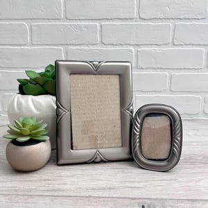 May include: Two silver-toned picture frames, one rectangular and one oval, are displayed on a light wood surface. The rectangular frame has decorative points at the corners. A small succulent plant in a concrete pot and a larger plant in a white pot are also present.
