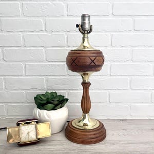 May include: A vintage table lamp with a wooden base and a brass accent. The lamp features a decorative wooden shade with an X pattern. A small, open travel clock and a potted succulent are also visible.