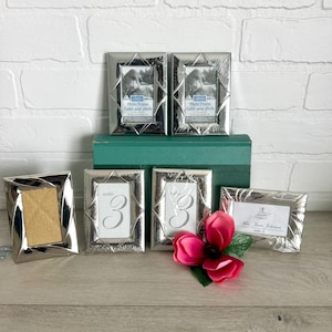 May include: A collection of six silver-toned metal photo frames with a geometric border design. Two frames contain inserts labeled Photo Frame 2.5 in x 3.5 in, two contain table number 3 cards, one is empty, and one holds a place card for Mrs. Jane Johnson.