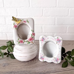 May include: Two white ceramic picture frames. One frame has a yellow bird perched on a branch with pink flowers. The other frame has a scalloped edge and is decorated with pink and blue floral ribbons. Both frames have a circular opening.