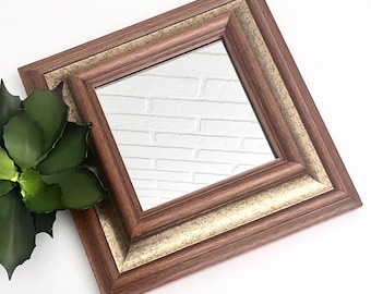 May include: A square mirror with a brown wooden frame and a gold inner trim. The mirror is reflecting a white brick wall.