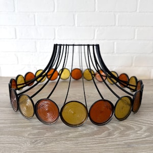 May include: A decorative, circular light fixture with a black metal frame. The fixture features a series of hanging, circular, amber, orange, and yellow glass discs. The discs are arranged around the perimeter of the fixture, creating a unique design.