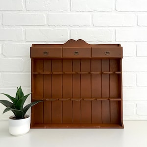 May include: A brown wooden wall-mounted shelf with three drawers and multiple slots. The shelf has a decorative curved top and brass drawer pulls.