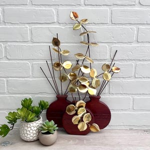 May include: A wall sculpture featuring three round, dark red vases with gold leaf accents. The vases are decorated with a geometric pattern and hold gold metal leaves.