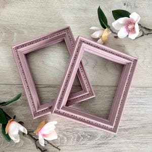 May include: Two rectangular picture frames in a muted mauve color, featuring a decorative border with a repeating leaf-like pattern. The frames are positioned at an angle, overlapping each other, and are accented by white magnolia flowers.