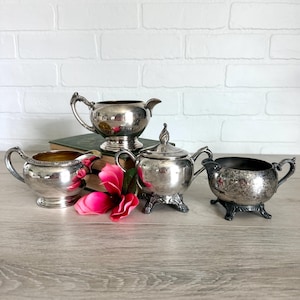 Puede incluir: Una colección de cuatro recipientes de metal plateado que incluye dos cremeras con asas, un azucarero con tapa y pies ornamentados, y un cuenco de boca ancha con acabado texturizado, dispuestos junto a una flor artificial de color rosa brillante.