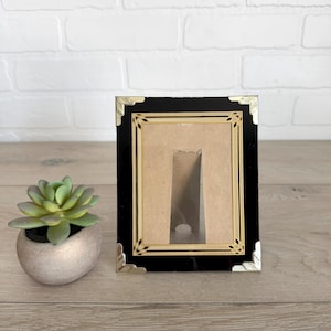 May include: A black picture frame with gold-colored corners and a beige interior. The frame is rectangular and has a glossy finish. A small succulent plant in a silver pot is to the left of the frame.