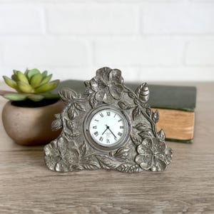 May include: A silver-toned metal desk clock featuring a decorative frame with embossed floral and leaf patterns. The clock face is white with black Roman numerals and thin black hands. The brand name Lenox is printed in the center of the clock face.