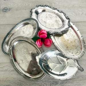 Puede incluir: Una colección de cinco bandejas de metal plateado de diversas formas ovaladas y de hoja, con bordes en relieve ornamentados y patrones grabados intrincados en las superficies, dispuestas con una flor artificial rosa brillante en el centro.