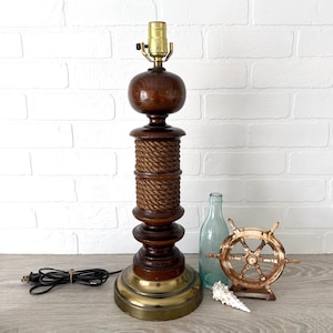 May include: A vintage table lamp with a dark wood base and a brass-colored bottom. The lamp features a rope-wrapped section and a bulb socket at the top. A nautical-themed decorative wheel and a blue glass bottle are next to the lamp.