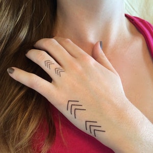 May include: A close-up of a person's hand with a simple black geometric tattoo of three sets of arrows pointing upwards on the back of their hand.