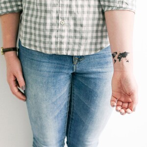 May include: A person wearing a gray and white plaid shirt and blue jeans. A black world map tattoo is on their wrist.