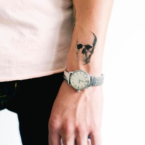 Może przedstawiać: Ramię osoby z tymczasowym tatuażem czaszki na przedramieniu. Osoba nosi srebrny zegarek z białą tarczą.