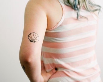May include: A black line art seashell temporary tattoo is displayed on an arm. The arm is in front of a person wearing a pink and white striped tank top. The tattoo is a simple outline of a seashell.