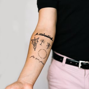 May include: Black ink tattoos on an arm featuring various designs, including a mountain range, a tree, a symbol, a ring, and the phrase "fly you fools". The tattoos are detailed and appear to be inspired by a fantasy theme.