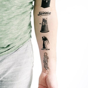 Op de afbeelding: Een arm met verschillende zwart-witte tijdelijke tatoeages geïnspireerd door de tv-serie Doctor Who. De tatoeages omvatten een TARDIS, een Dalek, een Weeping Angel en de TARDIS-consolekamer.