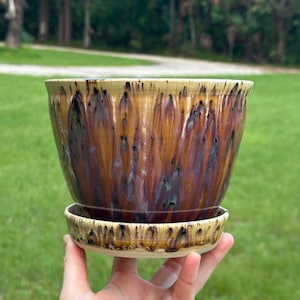 May include: A ceramic plant pot with a matching saucer featuring a glossy glaze in shades of amber, deep purple, and black. The surface displays a vertical drip pattern with a smooth finish, and the rim of the pot and saucer are a light tan color.