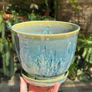 May include: A ceramic plant pot with a matching saucer, featuring a glossy glaze in shades of teal, blue, and green with a yellow-toned rim. The surface has a mottled, drip-like texture and a wide, circular opening at the top.