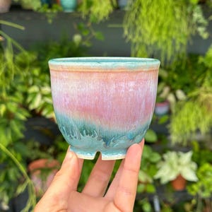 May include: A small, handmade ceramic planter with a unique glaze. The planter features a gradient of colors, including pink, blue, and green. The rim and base are a lighter shade of blue. The planter is designed for small plants and succulents.