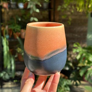 May include: A ceramic tumbler featuring a two-tone design with a matte peach-colored upper section and a dark charcoal-gray lower section. The vessel has a rounded, handleless silhouette with a smooth, slightly textured finish throughout.