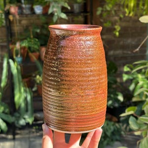 May include: A cylindrical ceramic vase featuring a textured, ribbed surface with a gradient glaze transitioning from deep brown at the base to a warm, speckled reddish-orange hue toward the flared rim, showcasing a handcrafted aesthetic.