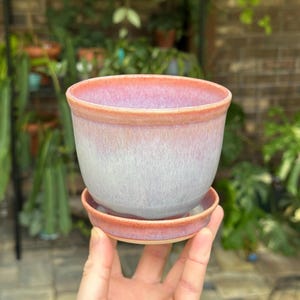 May include: A ceramic plant pot with a matching saucer, featuring a smooth glaze that transitions from a soft pink at the rim to a pale, muted lavender-gray at the base, with a distinct coral-toned edge on both the pot and the saucer.