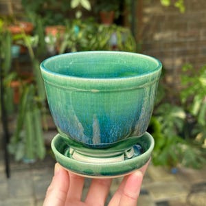 May include: A ceramic plant pot with a glossy glaze in shades of emerald green and deep teal. The pot features a rounded body, a wide rim, and a matching circular saucer base. The glaze shows subtle variations in color and texture throughout.