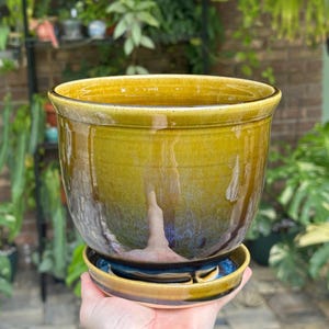 May include: A ceramic plant pot with a matching saucer featuring a glossy glaze that transitions from a warm olive green at the top to a deep, dark blue at the base. The pot has a wide, circular opening and a smooth, rounded rim.