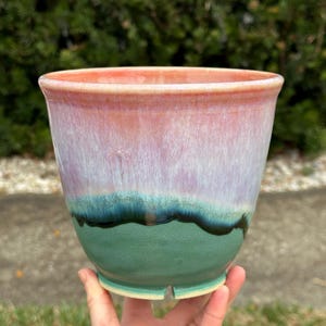 May include: A handmade ceramic planter with a gradient of colors. The top rim is a soft coral, transitioning to lavender, then teal, and finally a deep green base. The planter has a slightly flared shape.