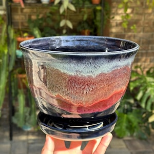 May include: A ceramic planter with a matching saucer featuring a glossy glaze that transitions from deep navy blue at the rim to a textured blend of light blue, cream, and vibrant red tones across the body. The pot has a wide, flared opening.