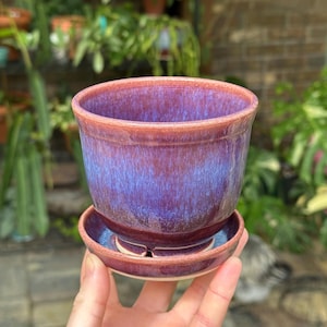 May include: A ceramic plant pot with a matching saucer, featuring a glossy glaze that transitions from a deep purple at the base to a lighter blue in the center, finished with a reddish-brown rim and a drainage hole at the bottom.