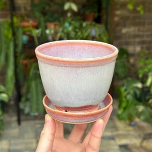 May include: A ceramic plant pot with a matching saucer, featuring a smooth, light gray body that transitions into a soft pink rim and base. The glaze has a subtle, blended finish, and the pot sits securely on the circular, lipped saucer.