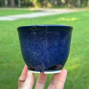 May include: A small, round ceramic bowl with a deep, glossy cobalt blue glaze featuring a speckled texture. The rim is a solid, dark navy blue, and the base is unglazed, revealing a light beige ceramic material with a small drainage notch.