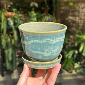 May include: A ceramic plant pot with a matching saucer, featuring a glossy glaze in shades of teal and blue with a textured, wavy white band around the center and a light green rim on both the pot and the saucer.