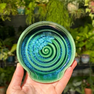 May include: A handmade ceramic spoon rest with a vibrant green and blue spiral design. The spoon rest has a circular shape with a small handle. The glaze creates a glossy, textured finish. Perfect for kitchen decor.