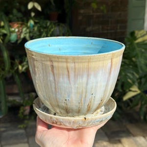 May include: A ceramic planter with a matching saucer, featuring a bright turquoise interior glaze and an exterior glaze with vertical drips of cream, tan, and brown tones over a smooth, rounded, bowl-shaped body.