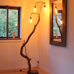 Op de afbeelding: Een unieke, handgemaakte vloerlamp gemaakt van een natuurlijke boomtak. De lamp heeft vier Edison-stijl gloeilampen, die een warme gloed geven. De houten basis en tak zijn bruin van kleur, wat een rustieke uitstraling geeft. De lamp is ongeveer 183 cm hoog.