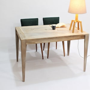 Puede incluir: Una mesa de madera clara con una lámpara de mesa de madera clara y dos sillas verde oscuro. La mesa tiene una tapa de madera clara y una base de madera clara. La lámpara tiene una base de madera clara y una pantalla beige clara. Las sillas tienen asientos de tela verde oscuro y patas de madera oscura.