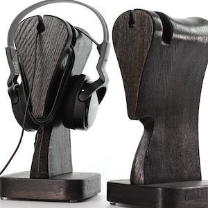 Op de afbeelding: Twee donkergrijze houten hoofdtelefoonstandaards. Eén standaard houdt een zwarte hoofdtelefoon vast. De standaards hebben een uniek, gebogen ontwerp met een sleuf om de hoofdtelefoon vast te houden. De basis van elke standaard heeft de tekst "PROJECT JK".