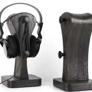 Puede incluir: Dos soportes de madera gris oscuro para auriculares. Un soporte sostiene auriculares negros con una diadema plateada. El otro soporte está vacío. Ambos soportes tienen una base cuadrada y un diseño curvo con perillas negras.