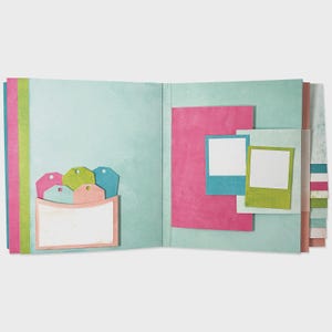 Puede incluir: Álbum de recortes abierto con páginas azul claro, con un bolsillo con etiquetas coloridas y marcos de fotos. Las páginas tienen detalles en rosa, verde y azul. Diseñado para fotos y diario.