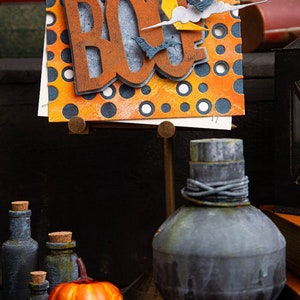 Può includere: Un biglietto a tema Halloween con la parola "BOO" in lettere arancioni e marroni su uno sfondo di pois arancioni e blu. Il biglietto è su un supporto con una piccola zucca e diverse bottiglie davanti.