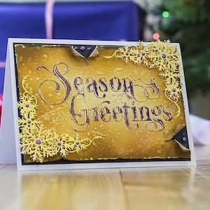 Pode incluir: Um cartão de felicitações artesanal com um fundo dourado e castanho e as palavras "Season's Greetings" em roxo. Recortes de flocos de neve dourados e detalhes roxos adornam os cantos. O cartão está sobre um fundo branco.