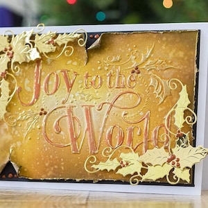Pode incluir: Um cartão de Natal dourado com o texto "Joy to the World" em uma fonte de escrita cursiva decorativa. O cartão é decorado com folhas de azevinho douradas e bagas.